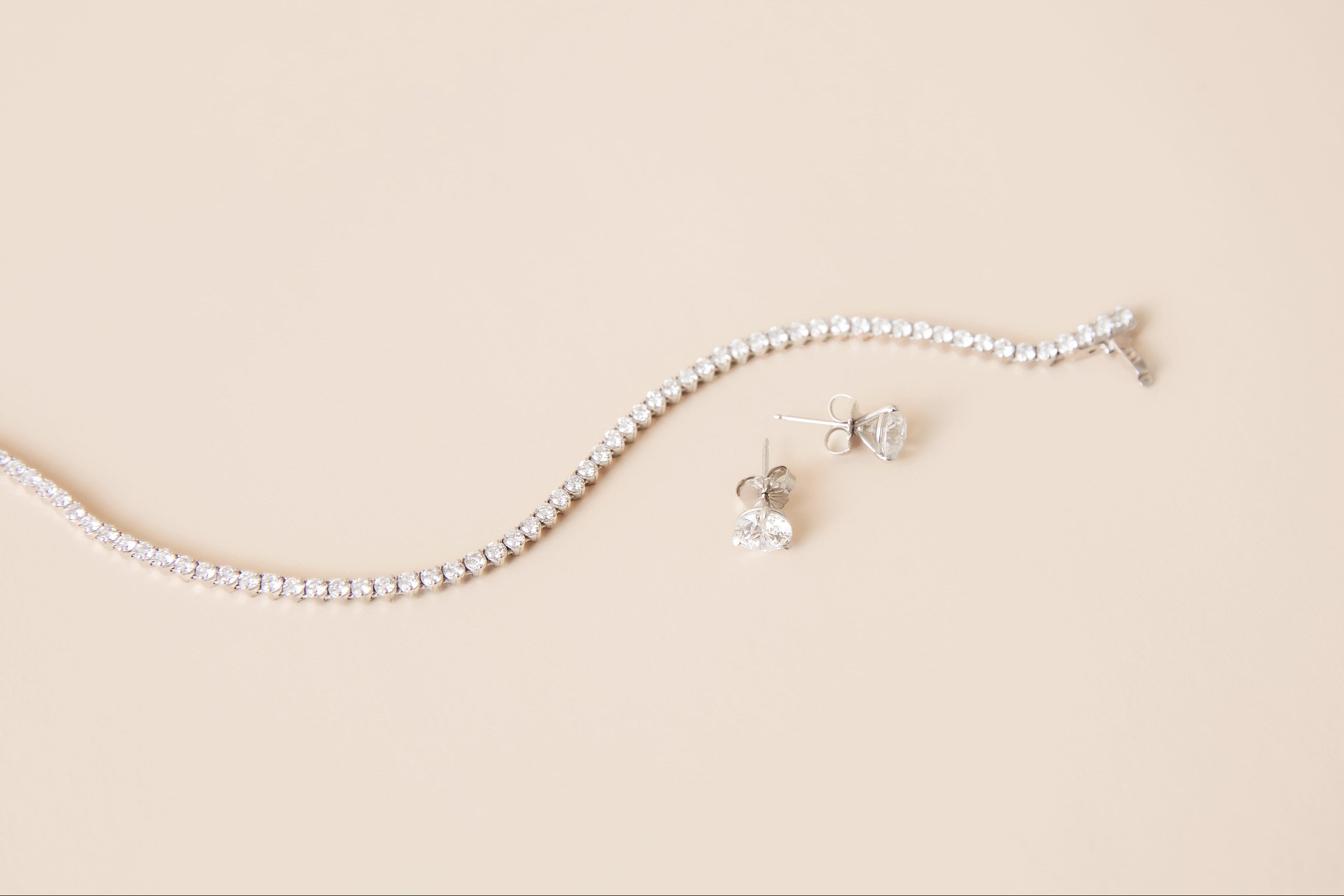 A white gold tennis necklace with diamonds and matching stud earrings, arranged on a solid beige background.