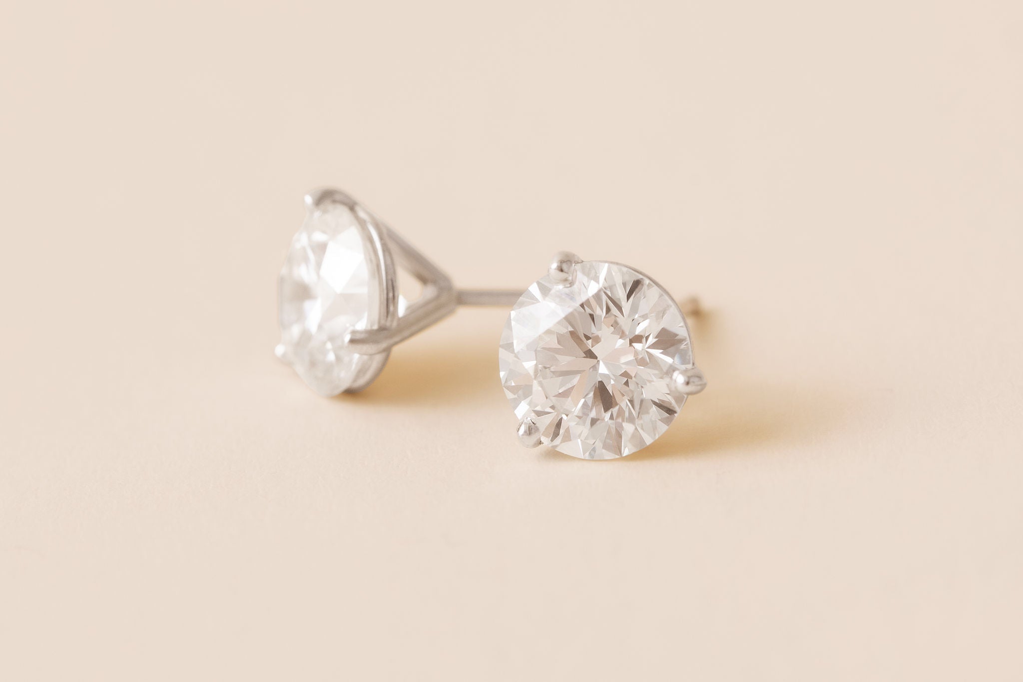 A pair of round-cut diamond stud earrings in a white gold setting, displayed on a soft beige background.