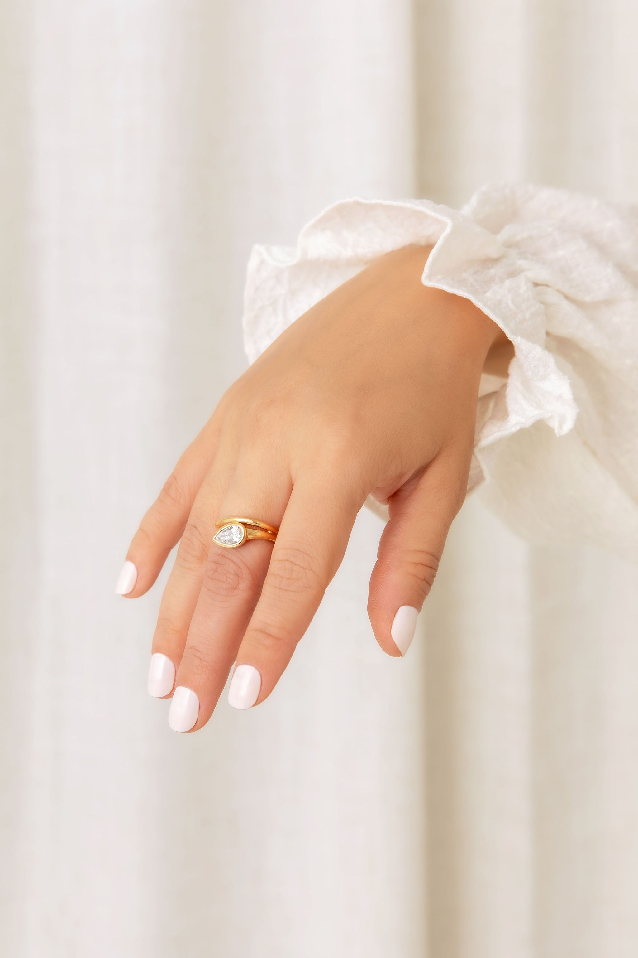 A gold ring with a pear-cut diamond worn on a hand with white manicured nails, styled with a white ruffled sleeve against a soft cream curtain background.