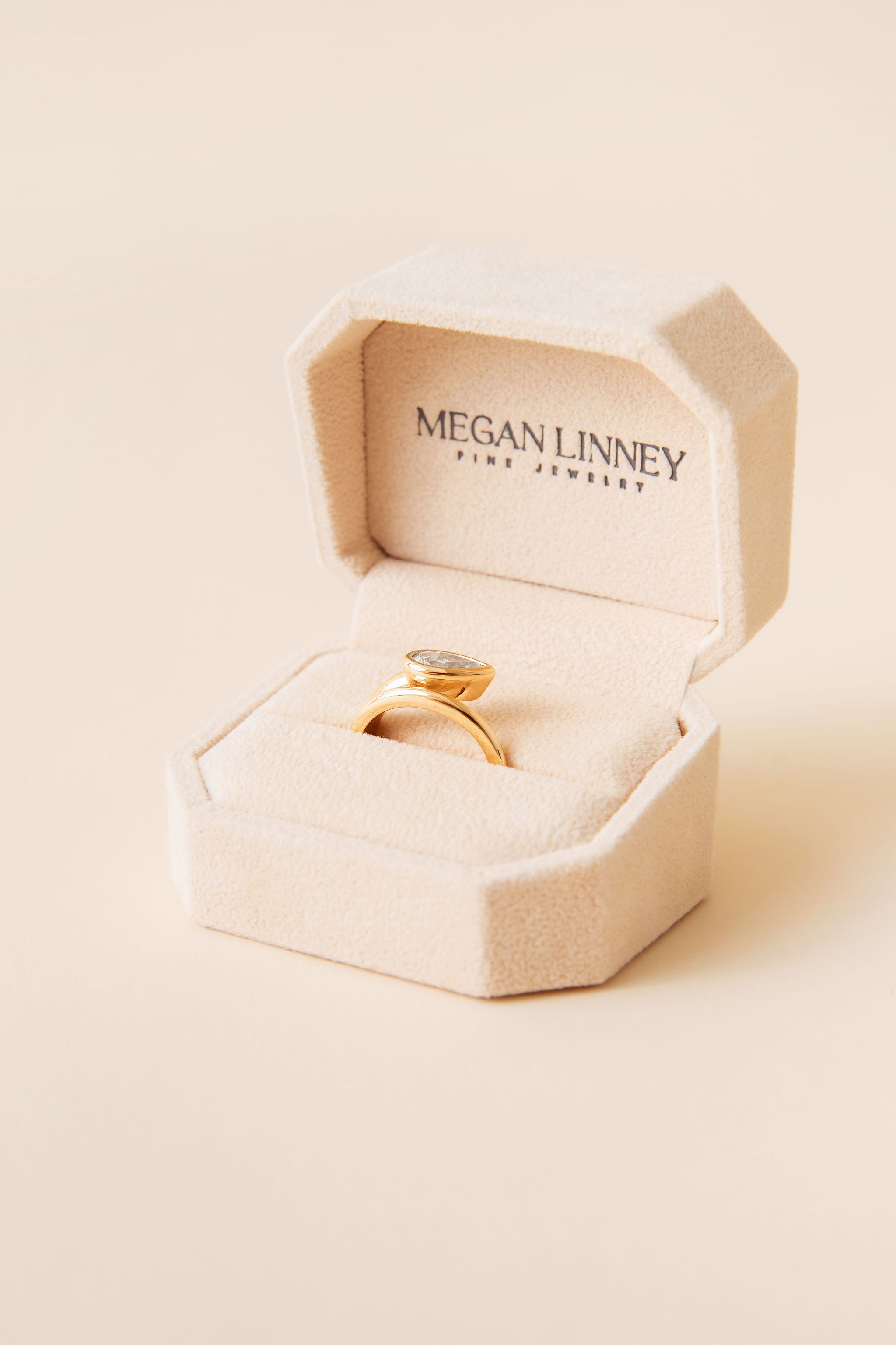 A gold ring with a pear-shaped diamond displayed in an open beige jewelry box. The inside lid of the box is printed with the brand name 'MEGAN LINNEY FINE JEWELRY'.