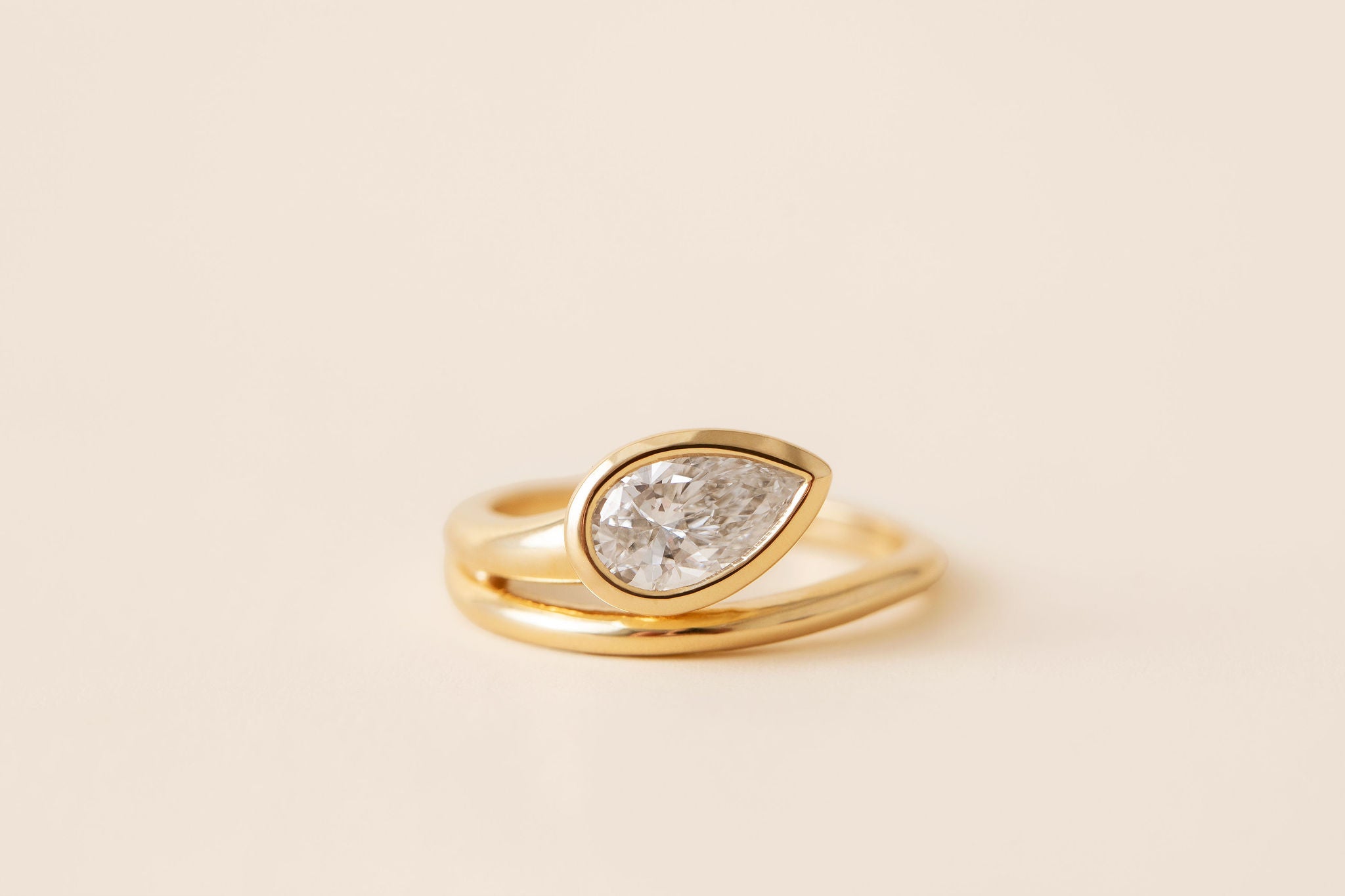 A gold ring featuring a pear-cut diamond in a bezel setting, with a second parallel gold band next to it, shown on a warm cream background.