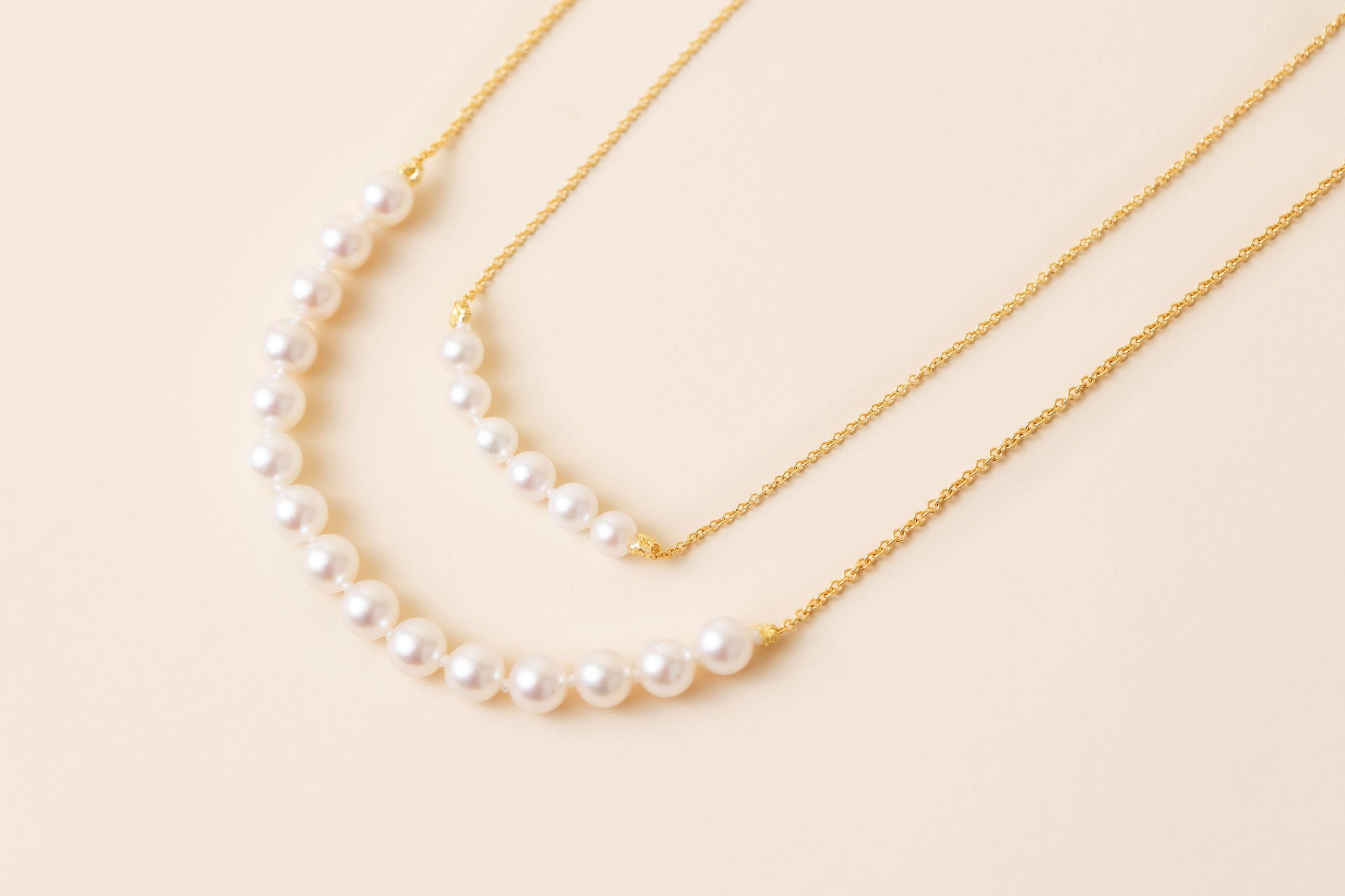 Two delicate gold chain necklaces with curved pearl strands, displayed on a soft beige background.