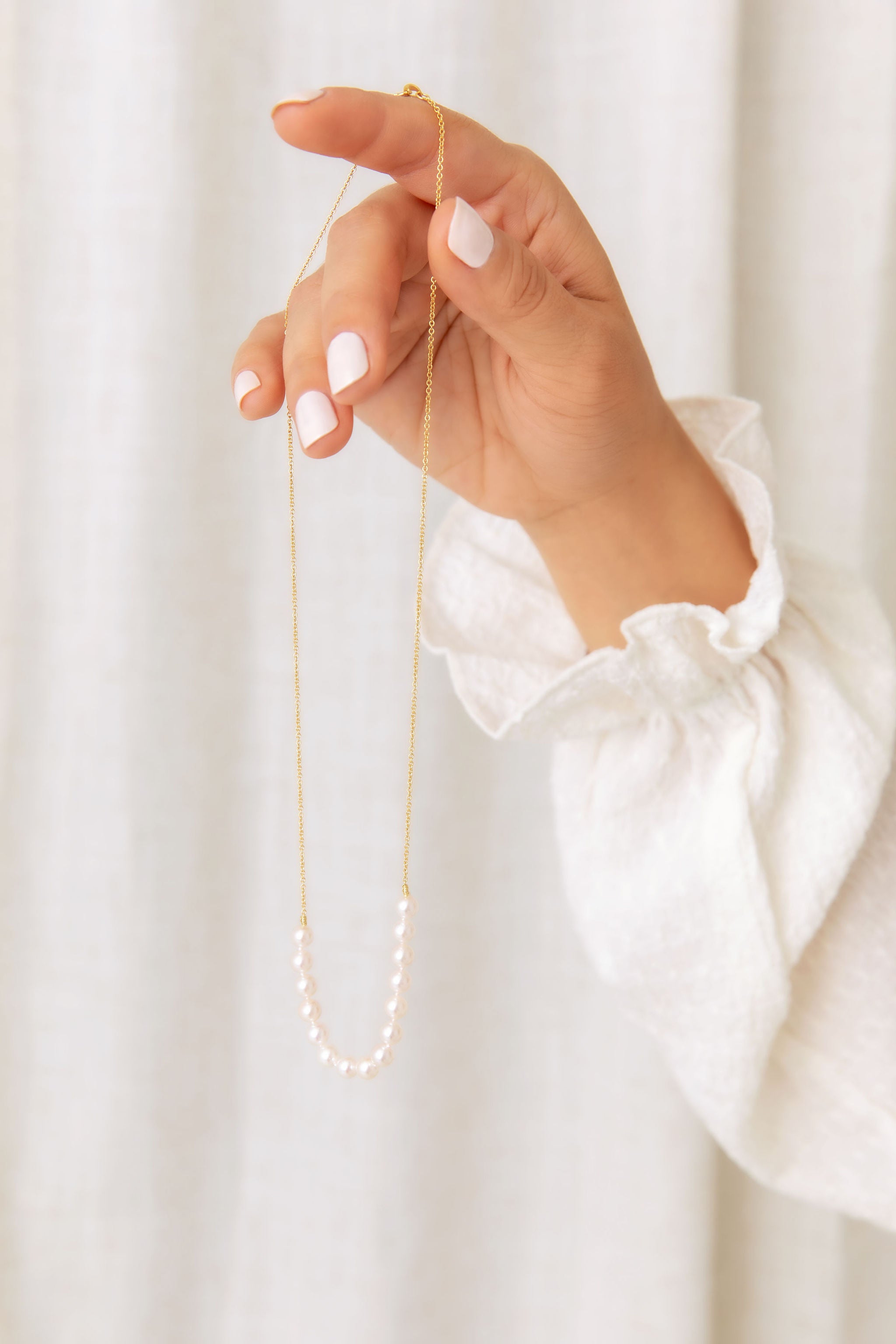 A close-up of a person's hand holding a delicate gold chain necklace with a curved band of white pearls, set against a soft white curtain background.