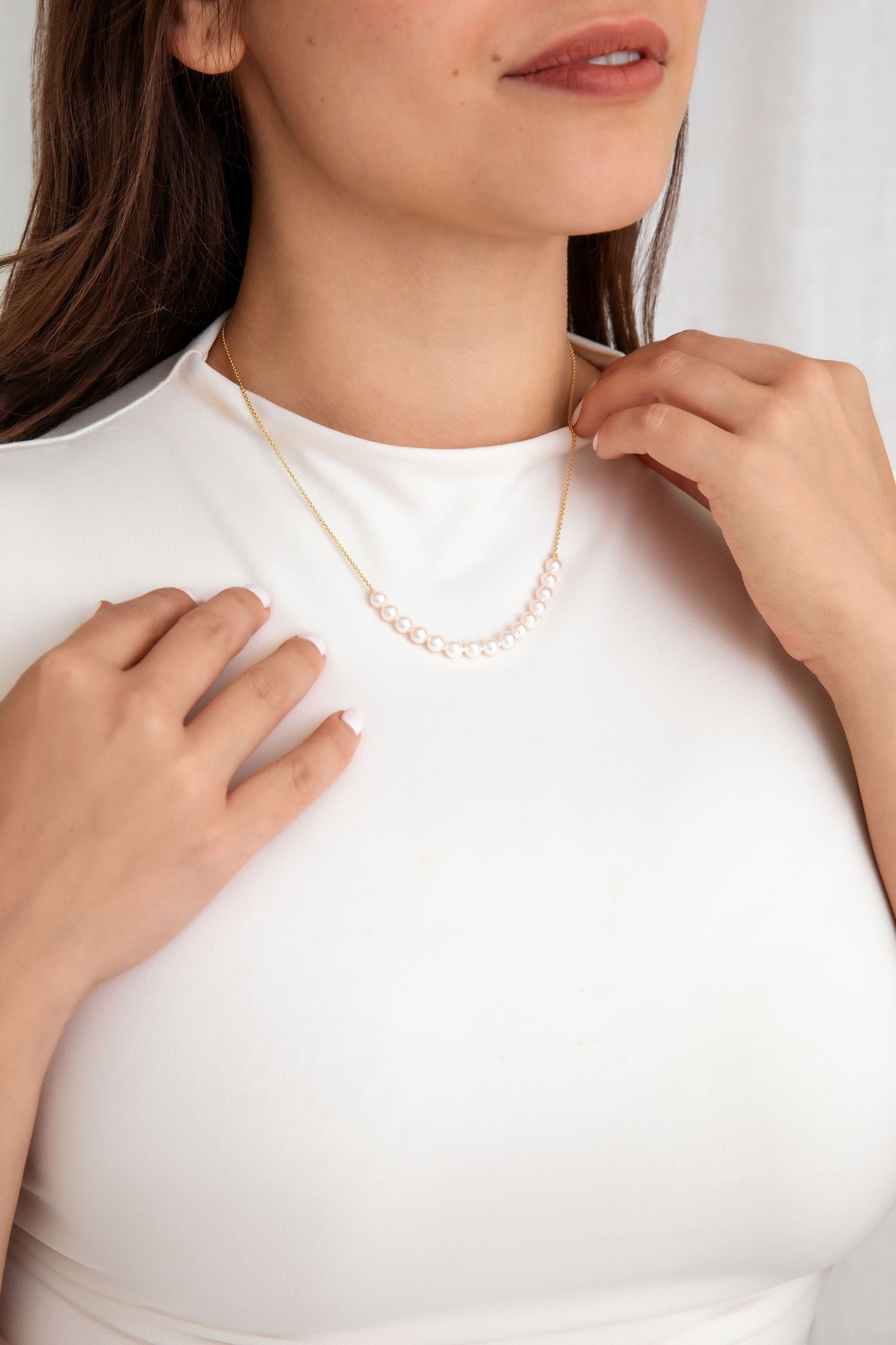 A close-up of a woman wearing a delicate gold necklace with a curved row of white pearls, styled with a white mock-neck top.