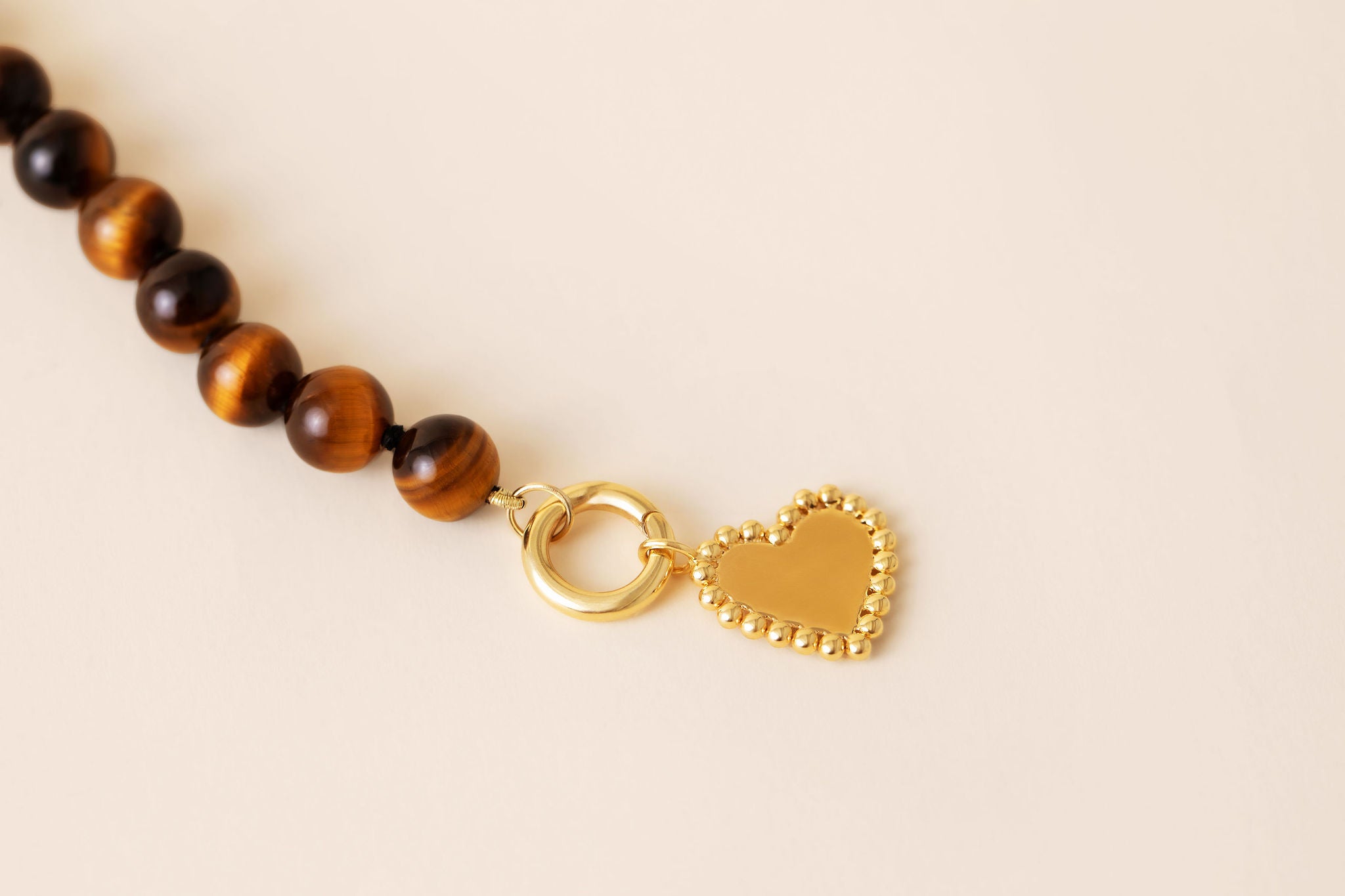 A close-up of a beaded necklace featuring round brown and black tiger's eye beads, connected by a gold ring to a small gold heart charm with a beaded border, set against a solid light beige background.