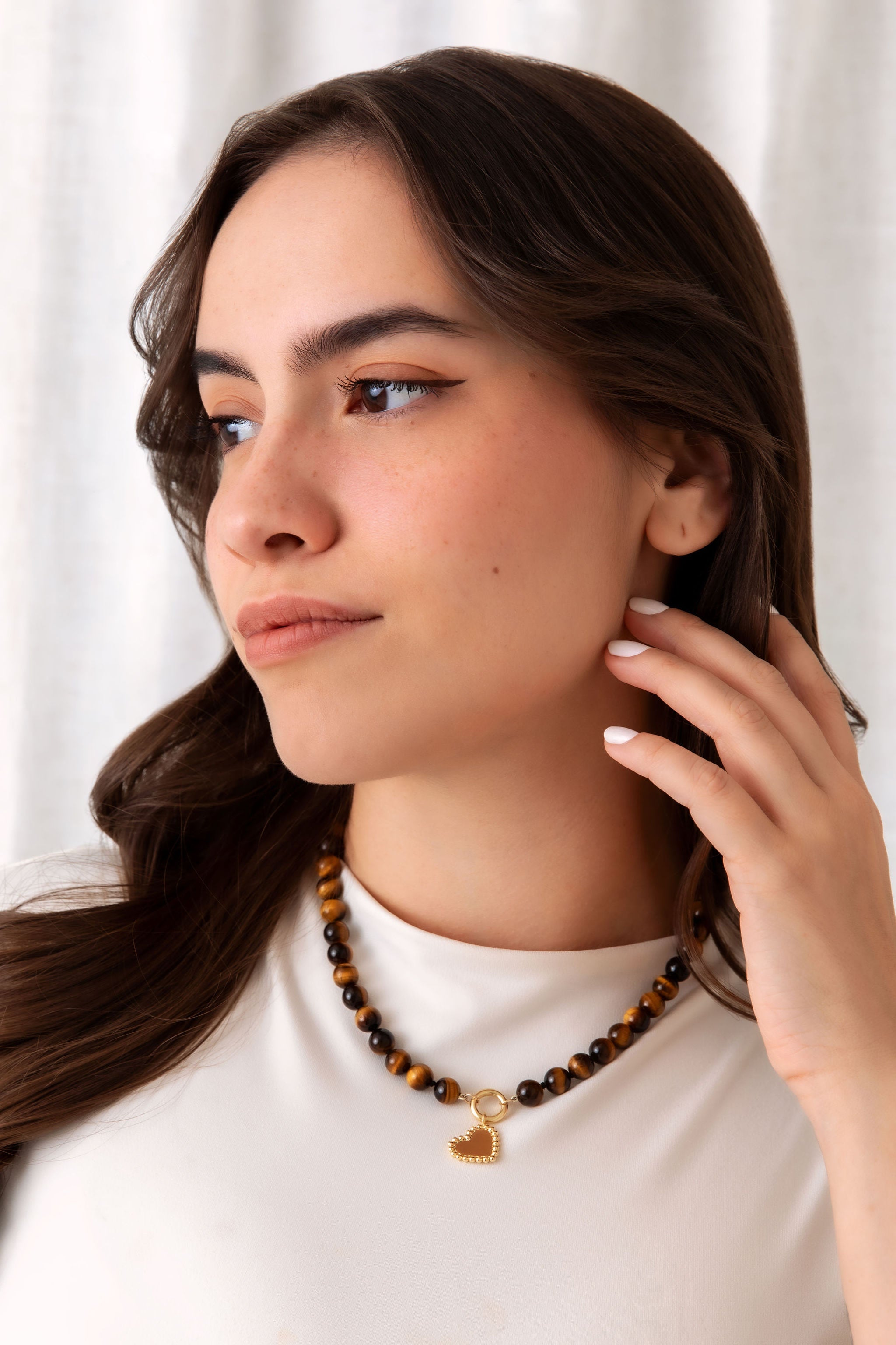 A woman with long dark brown hair wearing a beaded necklace with brown and black tiger's eye beads and a small gold heart charm, paired with a white top against a soft white curtain background.