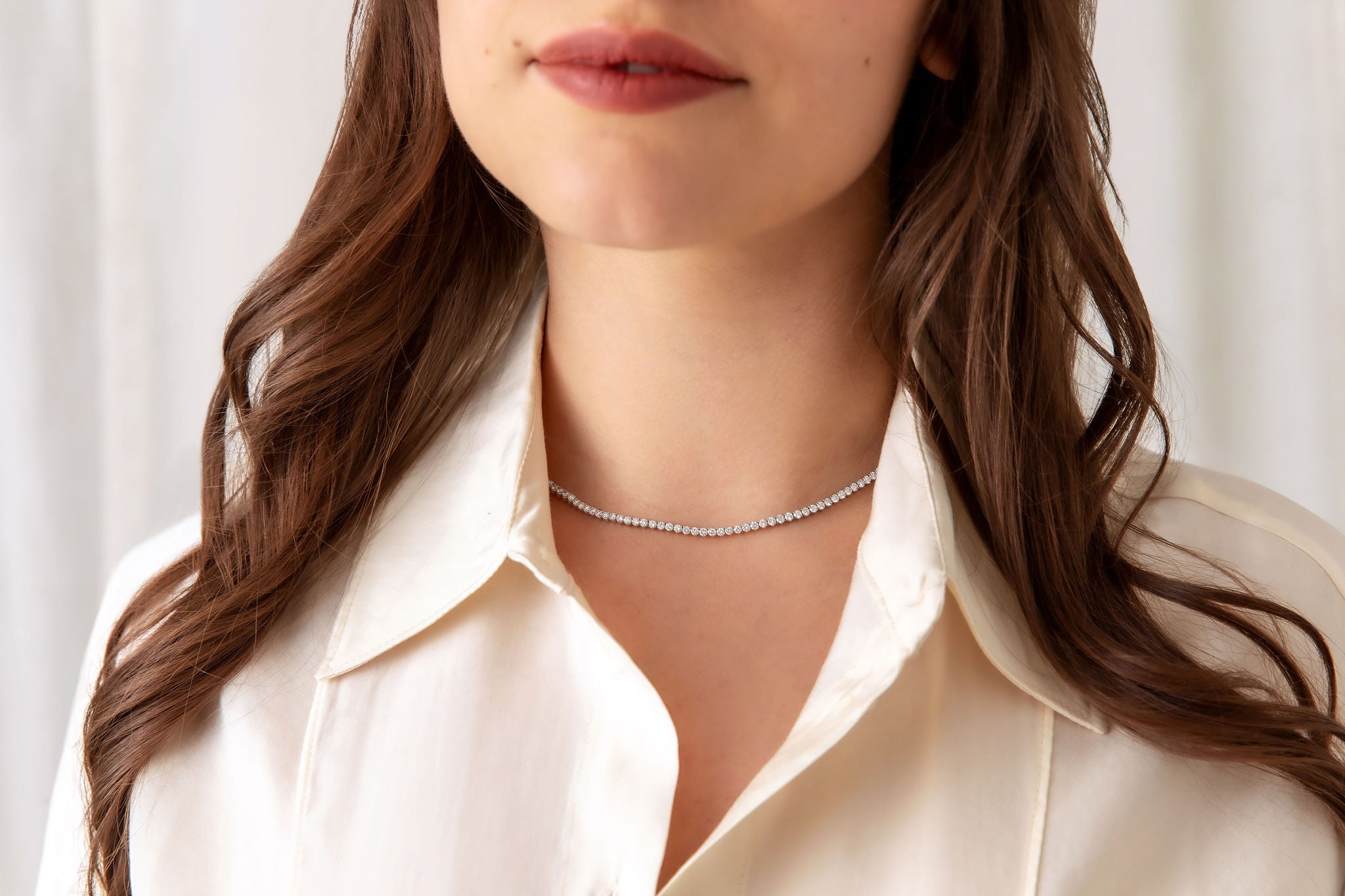 A close-up of a woman wearing a diamond tennis necklace, paired with a cream-colored collared shirt against a soft, light background.