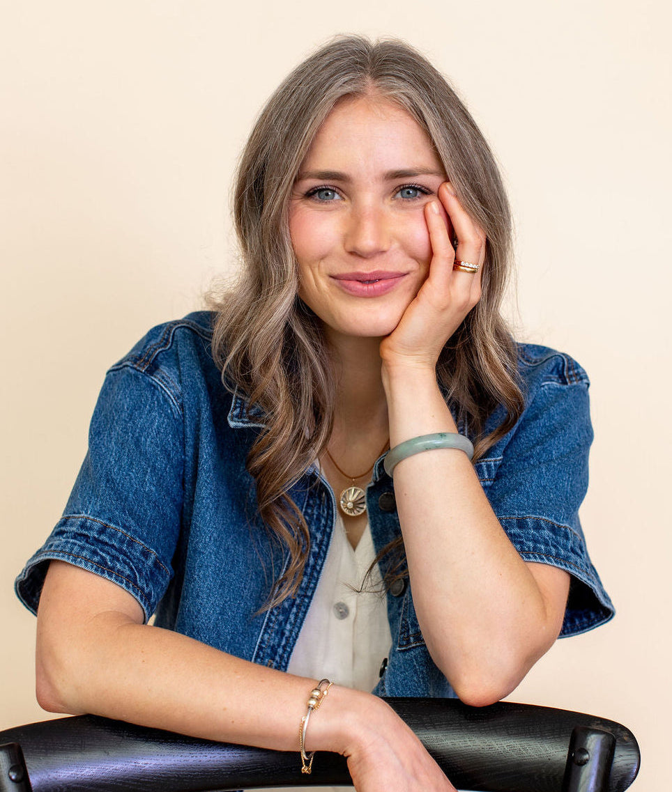 A woman with wavy light brown hair leans on a black chair, wearing a blue denim jacket over a white top. She is posed against a solid beige background.