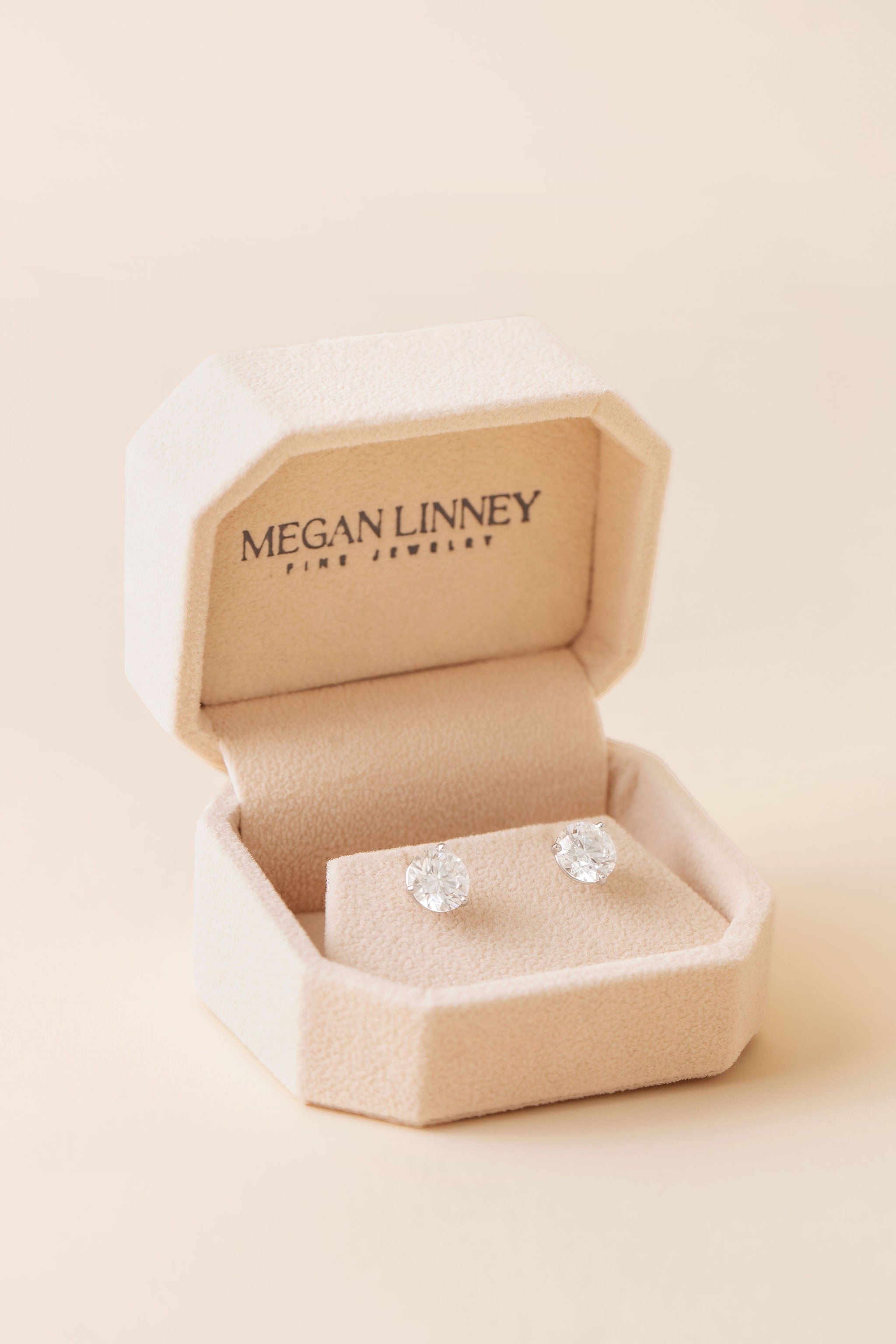 A pair of round-cut diamond stud earrings displayed on a beige velvet jewelry box lid, with the brand name 'MEGAN LINNEY FINE JEWELRY' printed inside the box against a warm cream background.