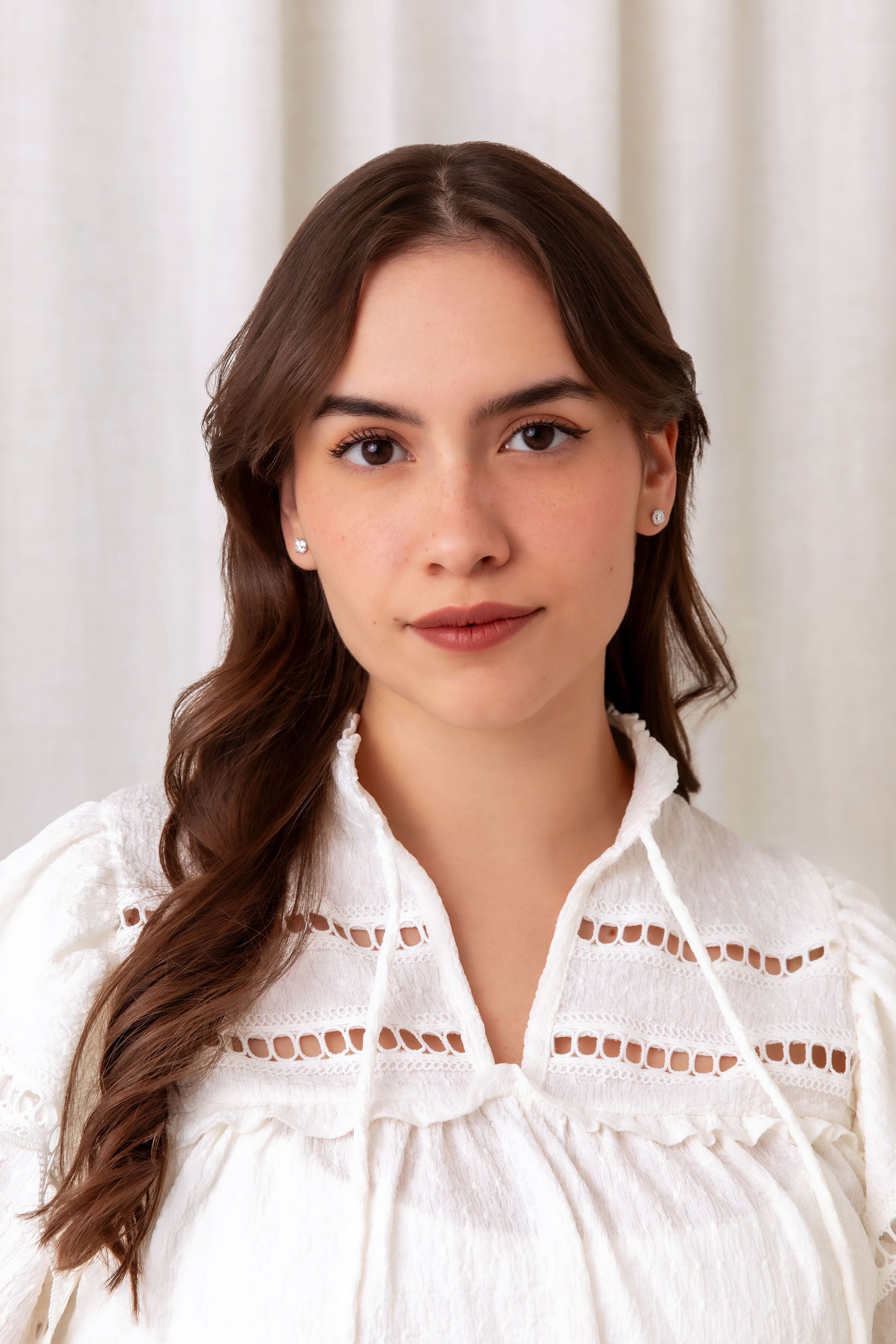 A woman with long brown hair wearing a white eyelet lace blouse and diamonds stud earrings, photographed against a soft off-white curtain background.