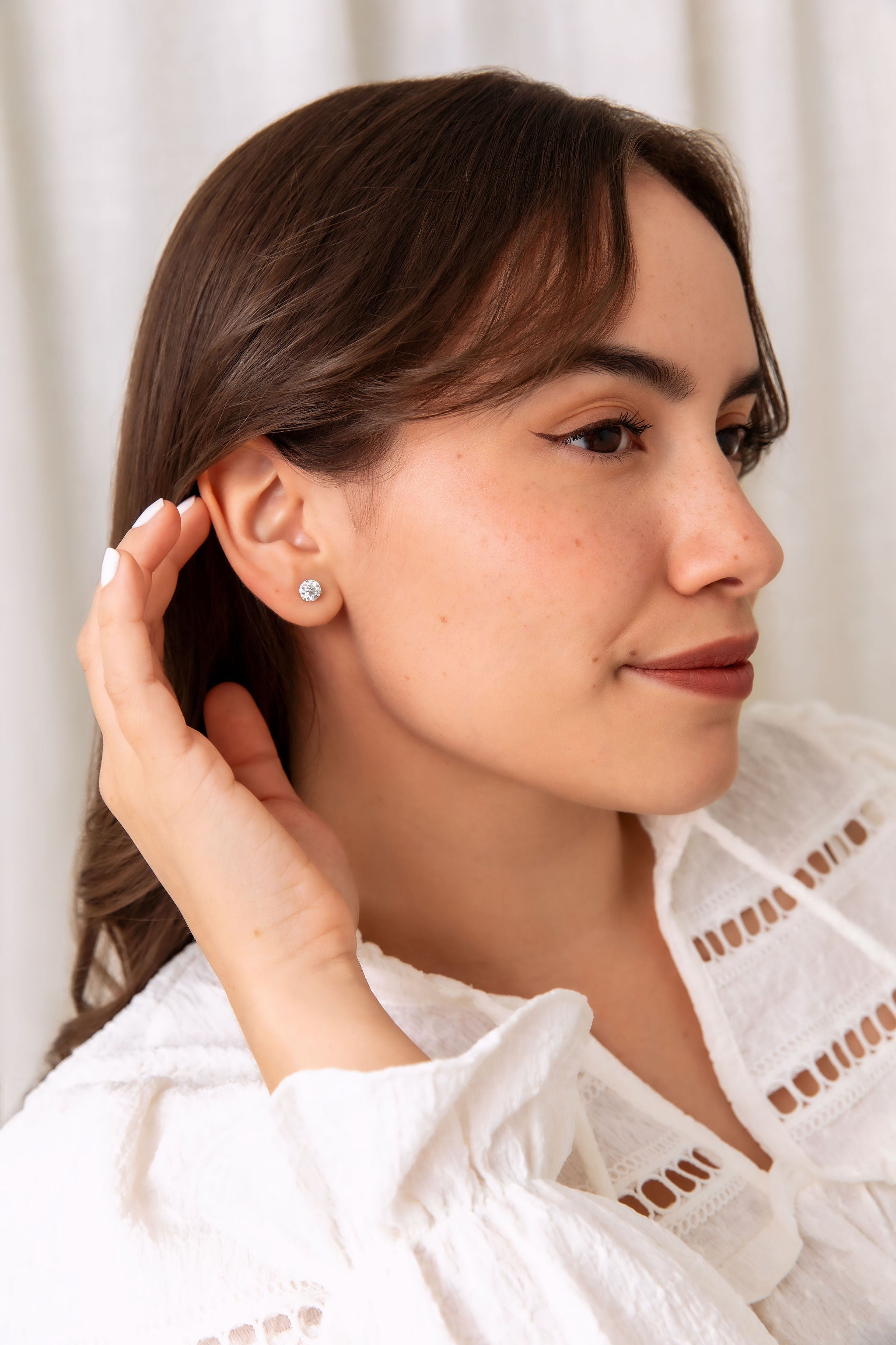 A woman in a white eyelet blouse wearing a small silver stud earring, shown in a close-up profile against a soft white curtain background.