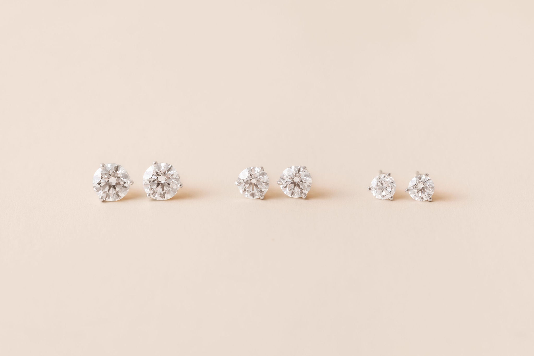 Three pairs of white gold stud earrings with diamonds, arranged in a row on a solid beige background.