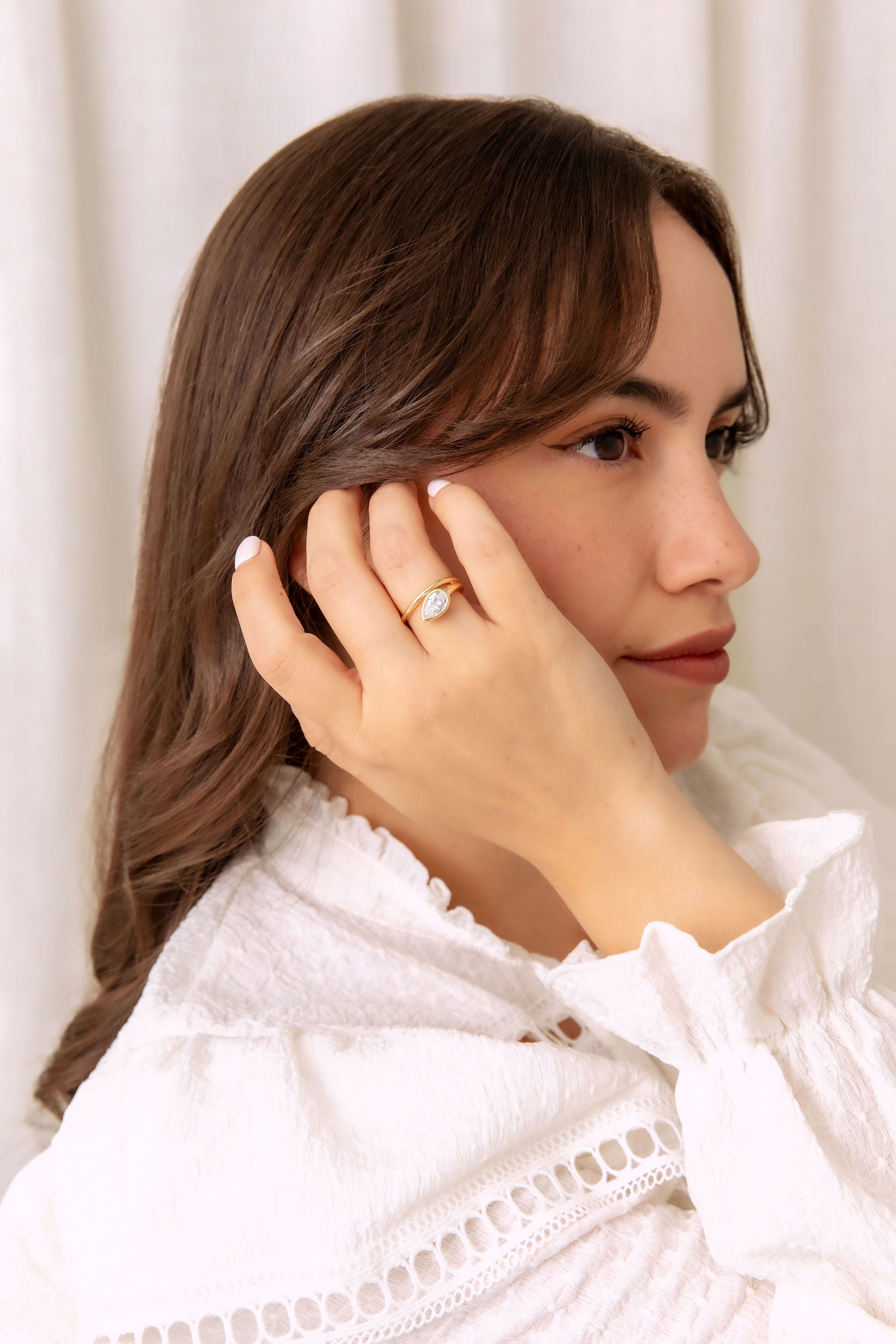 A woman with long brown hair wearing a white blouse, shown in profile with her hand near her face, wearing a gold wrap ring with a pear shaped diamond.
