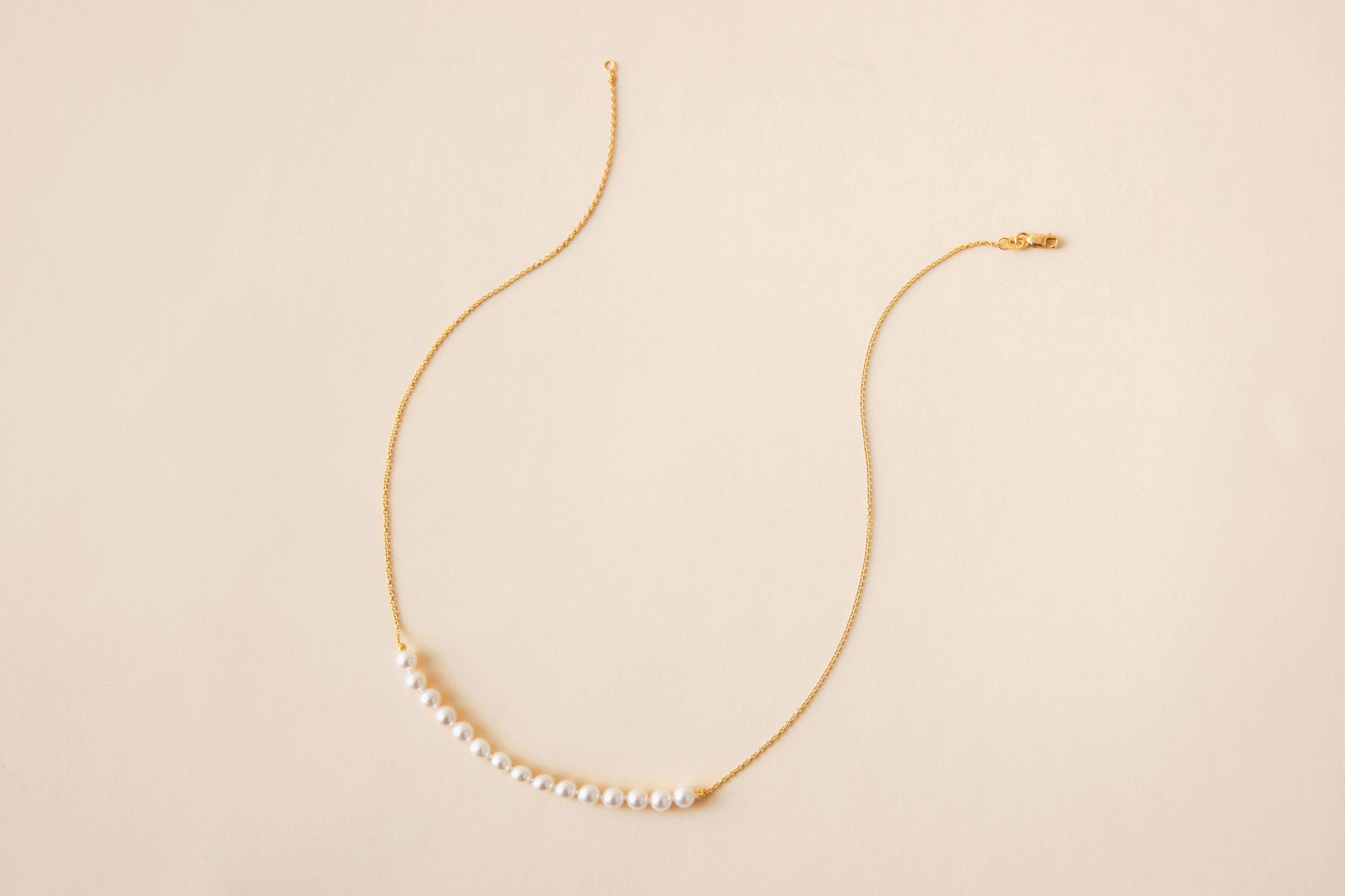 A delicate gold chain necklace featuring a curved bar of white pearls at the center, displayed on a solid light beige background.