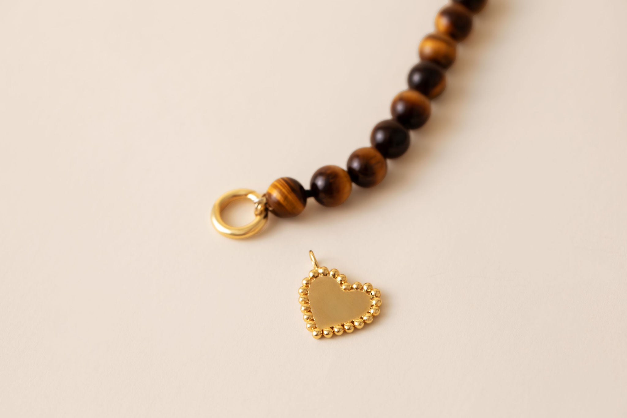 A close-up of a beaded necklace with dark brown and golden-brown round tiger's eye beads, a gold ring clasp, and a small gold heart-shaped charm on a light beige background.