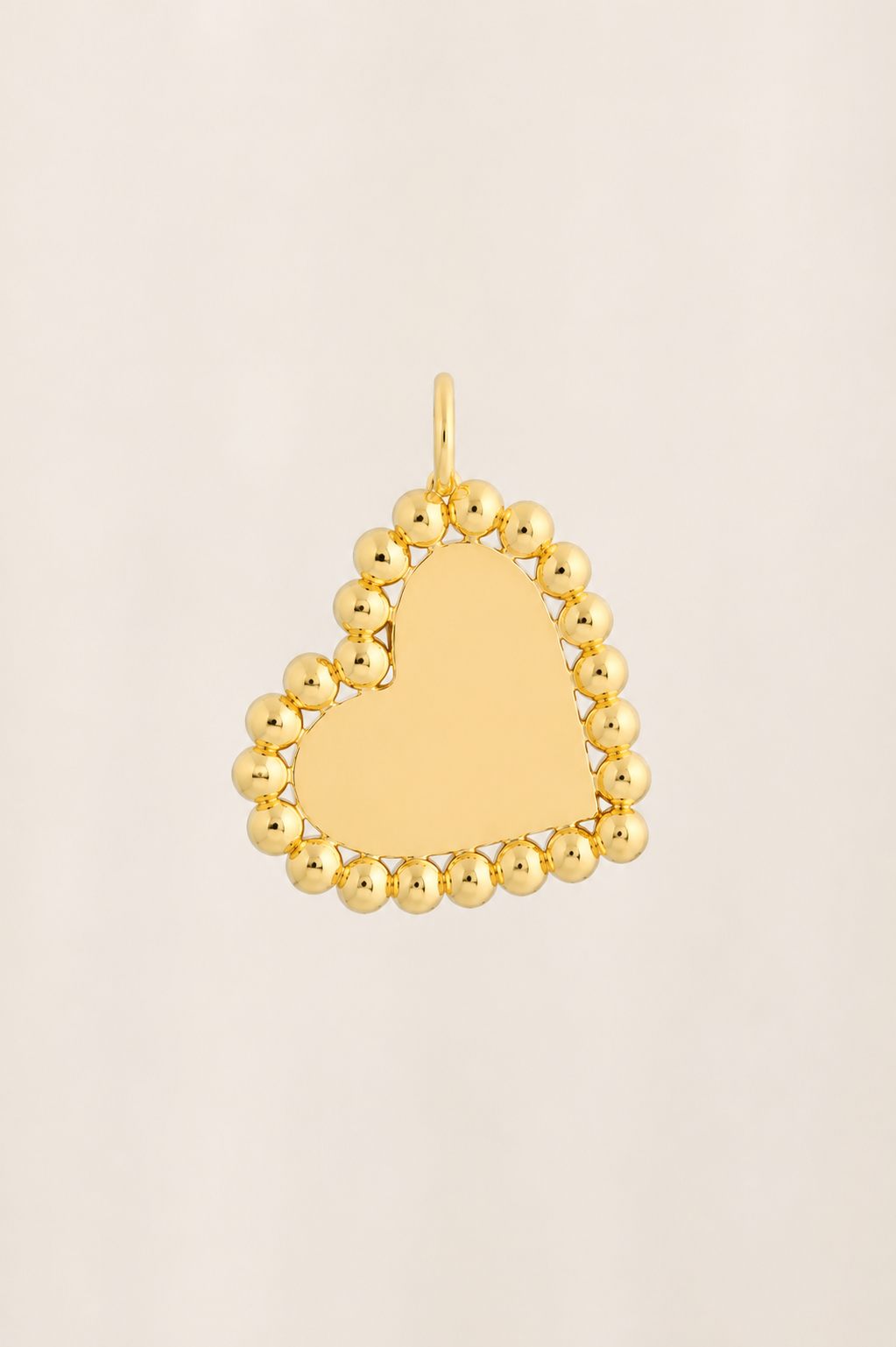 A gold heart-shaped pendant with a smooth, polished center and a border of small, rounded beads, shown on a light beige background.