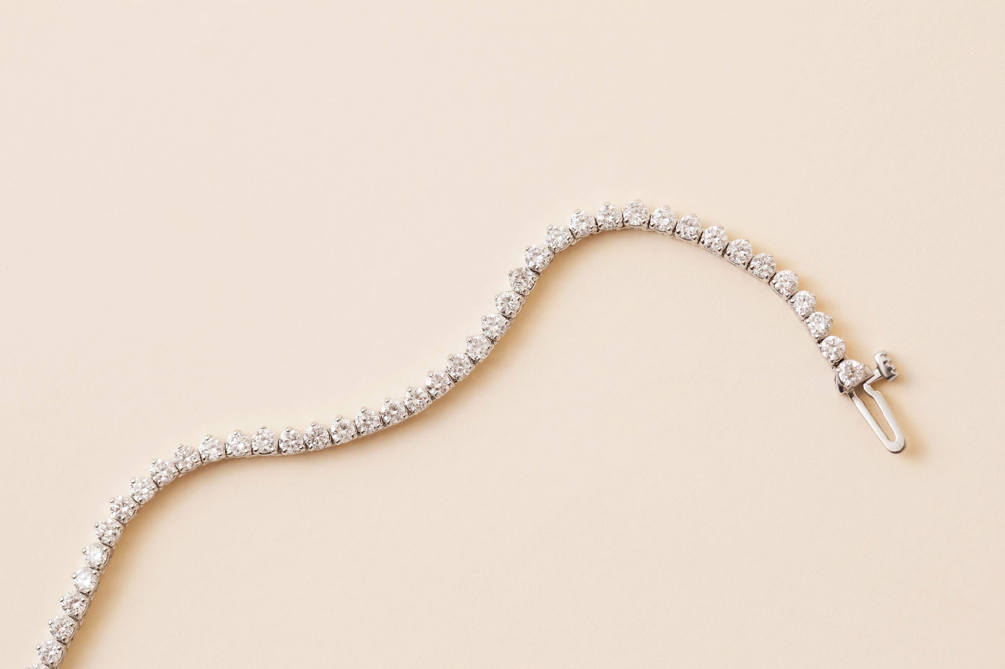 A diamond tennis necklace in white gold, arranged in a curved S-shape on a solid pale beige background.