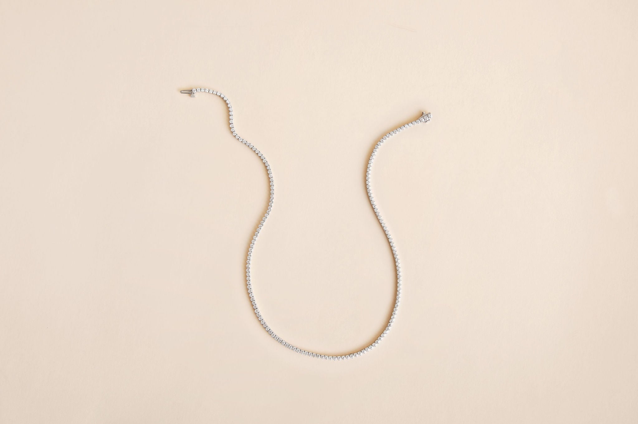 A delicate white gold diamond tennis necklace arranged in a U-shape on a solid, warm beige background.