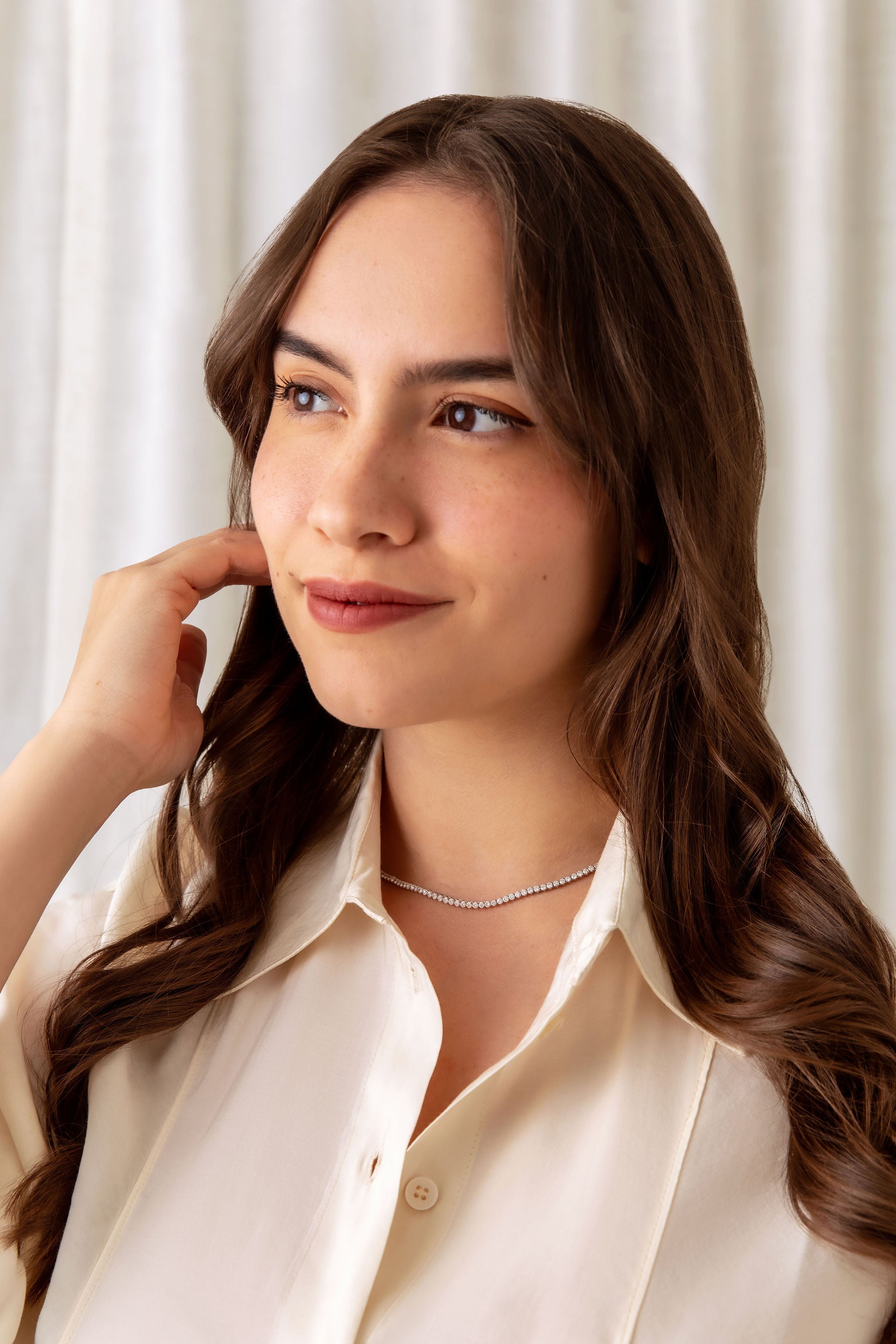 A woman with long brown hair wearing a delicate diamond tennis necklace and a cream-colored collared shirt, posing against a soft, light beige curtain background.