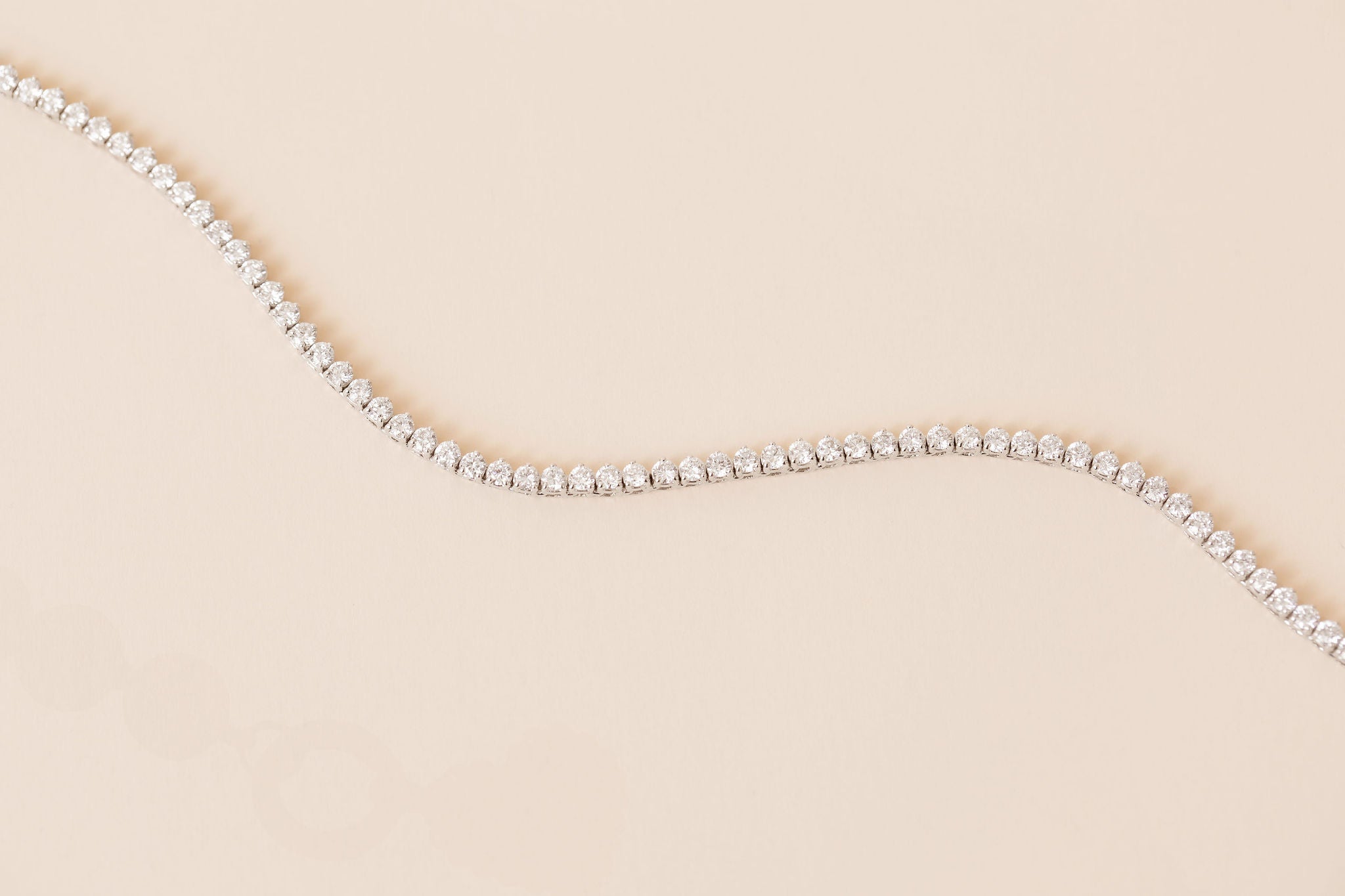 A delicate white gold diamond tennis necklace, arranged in a curved line on a solid pale peach-beige background.